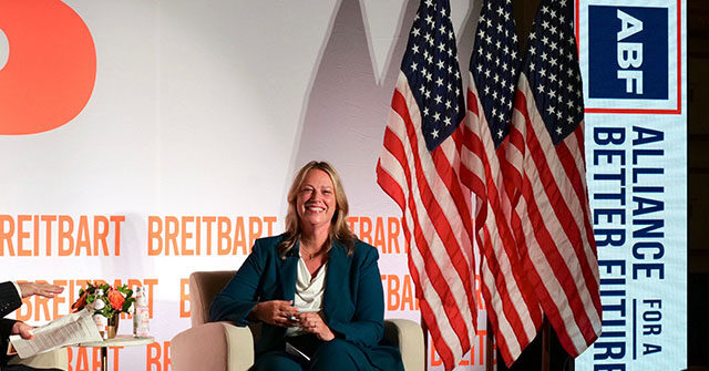 Janet Kelly seated on stage during a Breitbart interview, with American flags and an Alliance for a Better Future backdrop